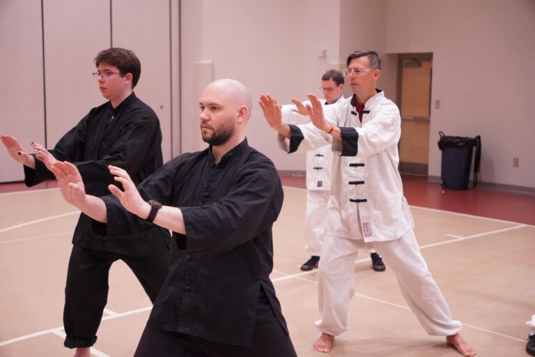 Tai Chi students in black and white uniforms practicing synchronized stance in training hall